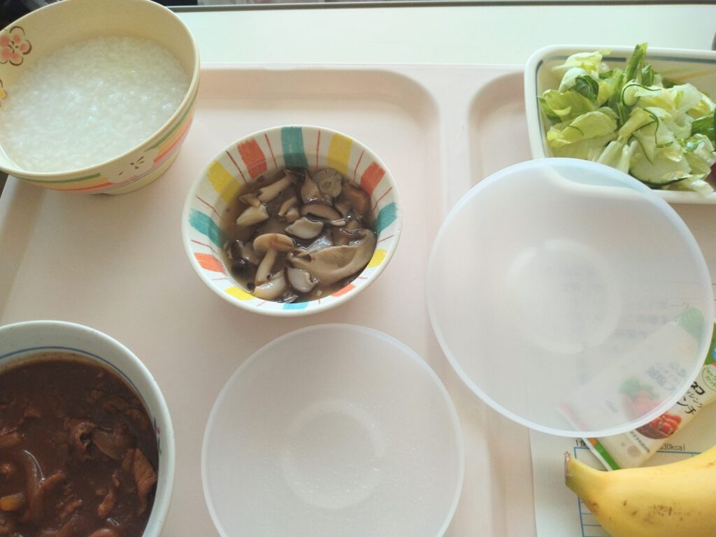 子宮全摘手術の翌日に出された病院の昼食。お粥、ビーフシチュー、和え物、バナナ、ジョアがトレイに並んでいる写真。